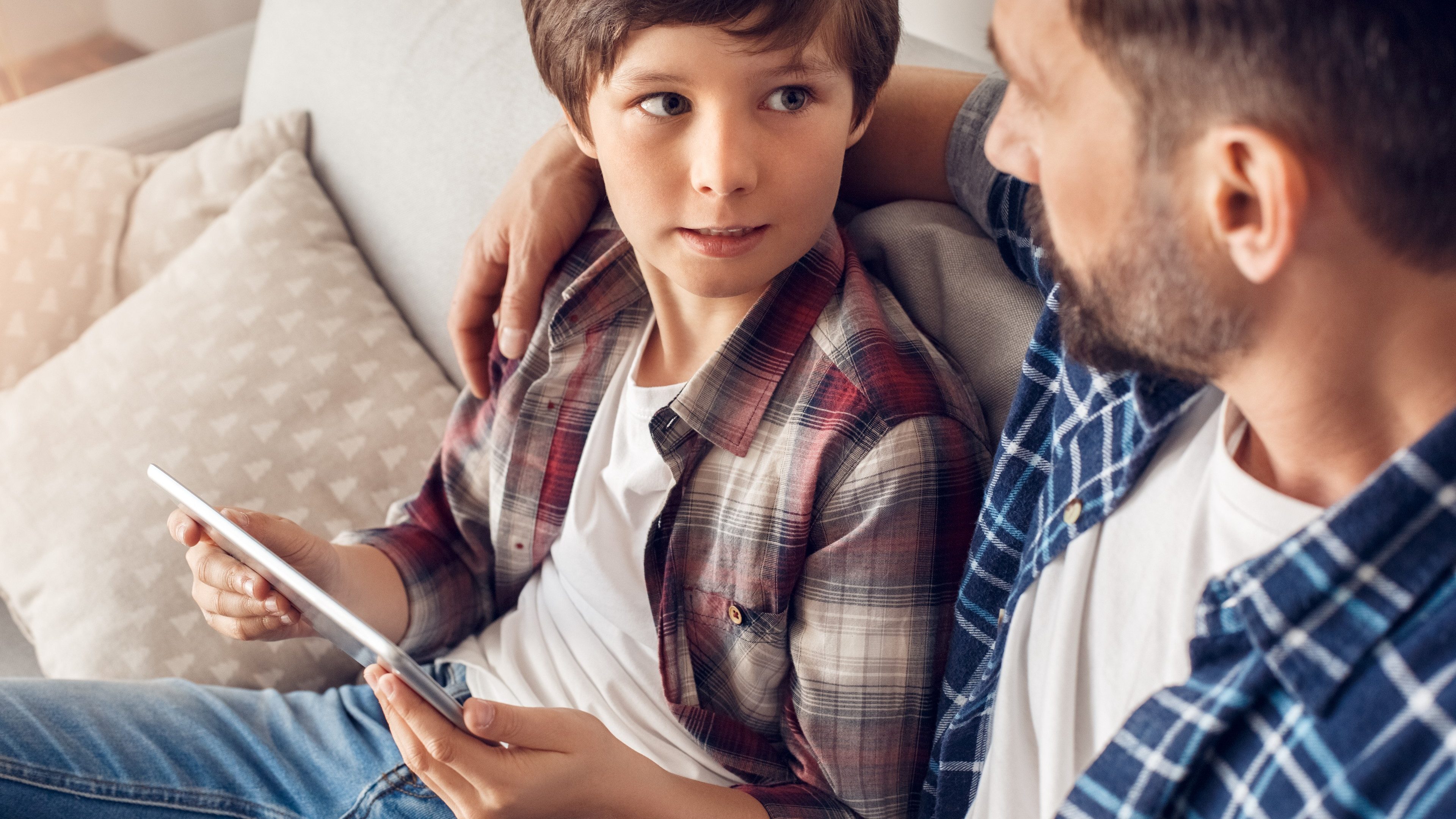 Father and little son together at home sitting on sofa dad hugging boy holding digital tablet looking at each other serious close-up