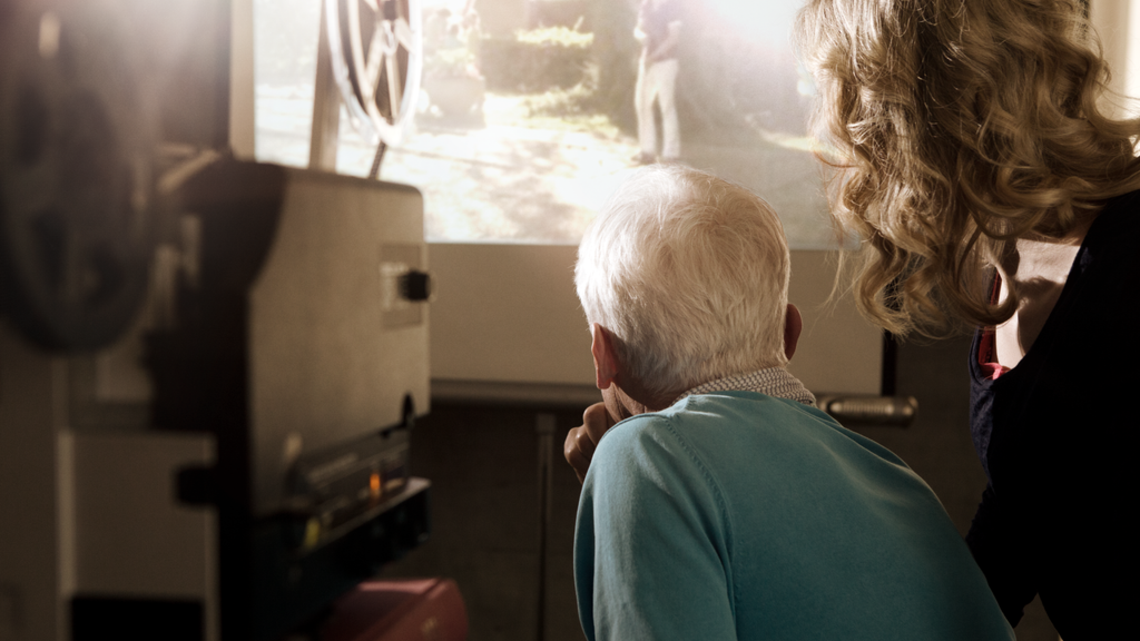 Un monsieur âgé et une jeune femme, de dos, regardent un film en format super 8 mmm sur un écran tendu