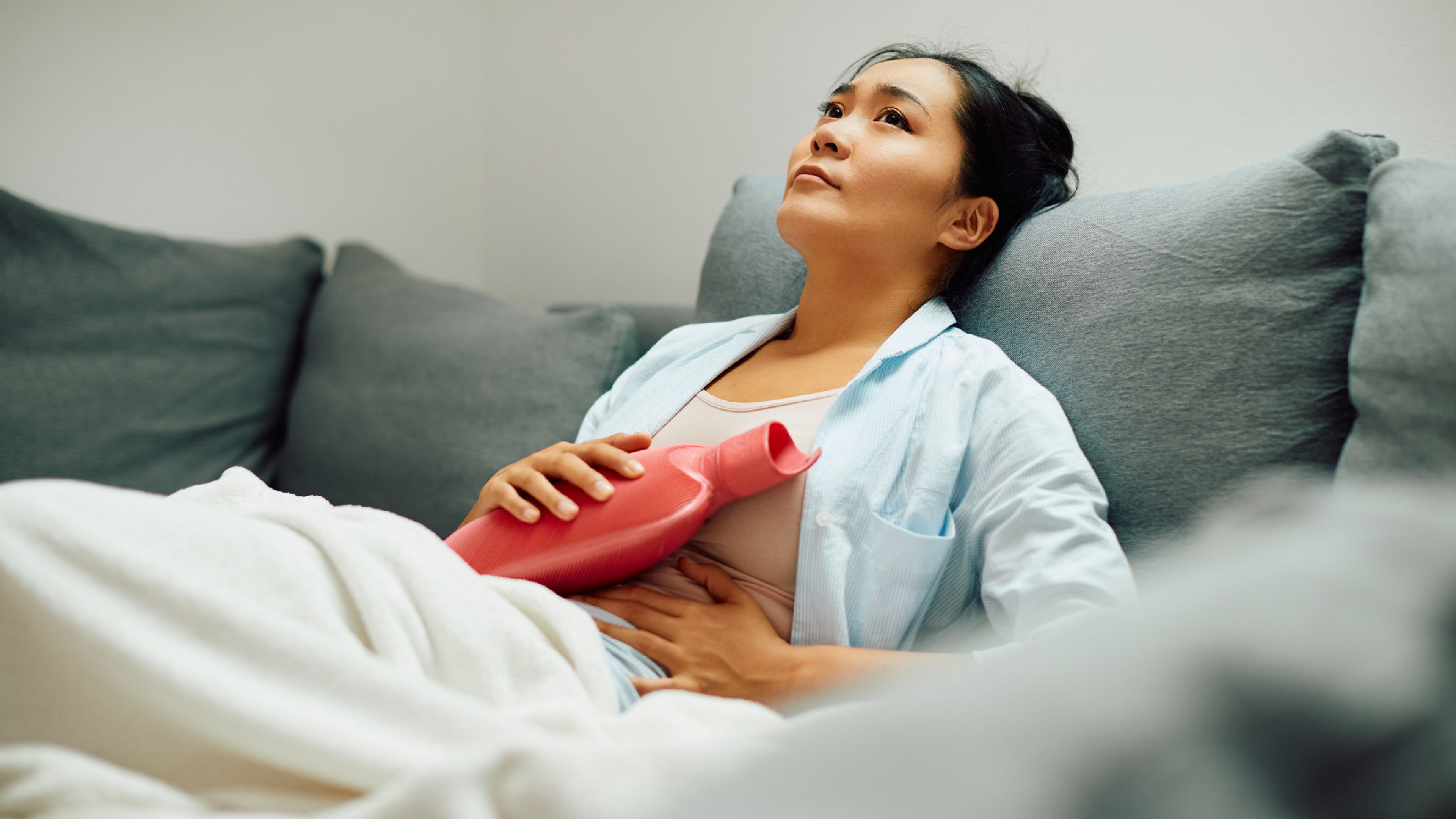 Young Asian woman relieves stomach pain with hot water bottle while relaxing on sofa at home.