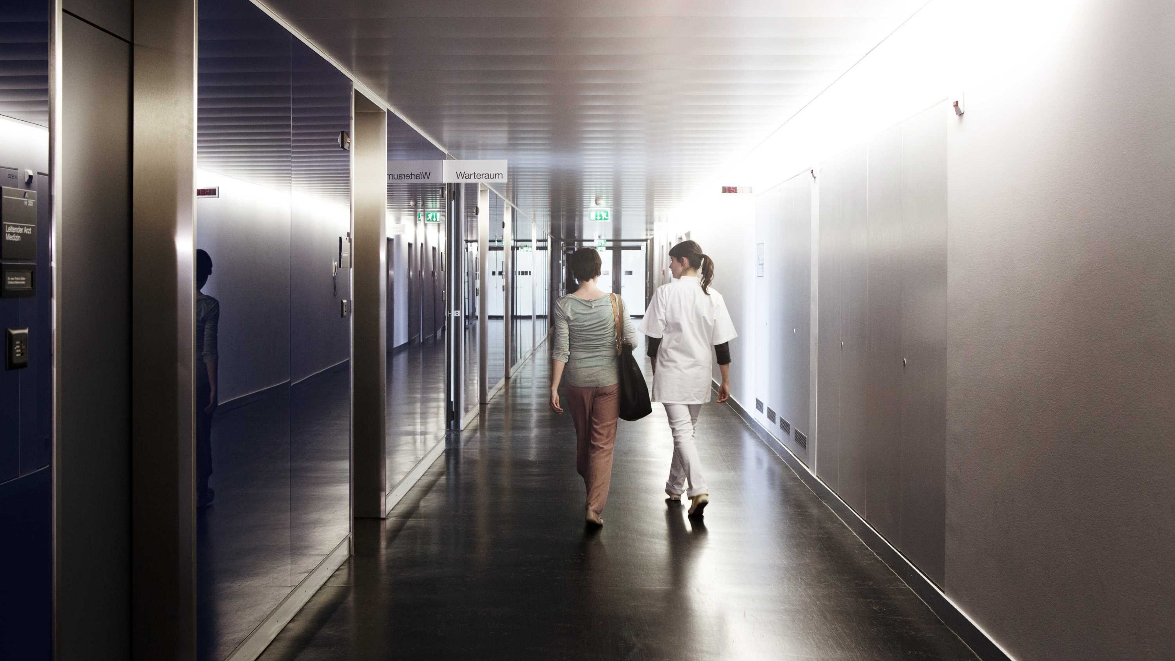 Deux femmes de dos dont une en blouse blanche qui marchent dans les couloirs d'un hopital