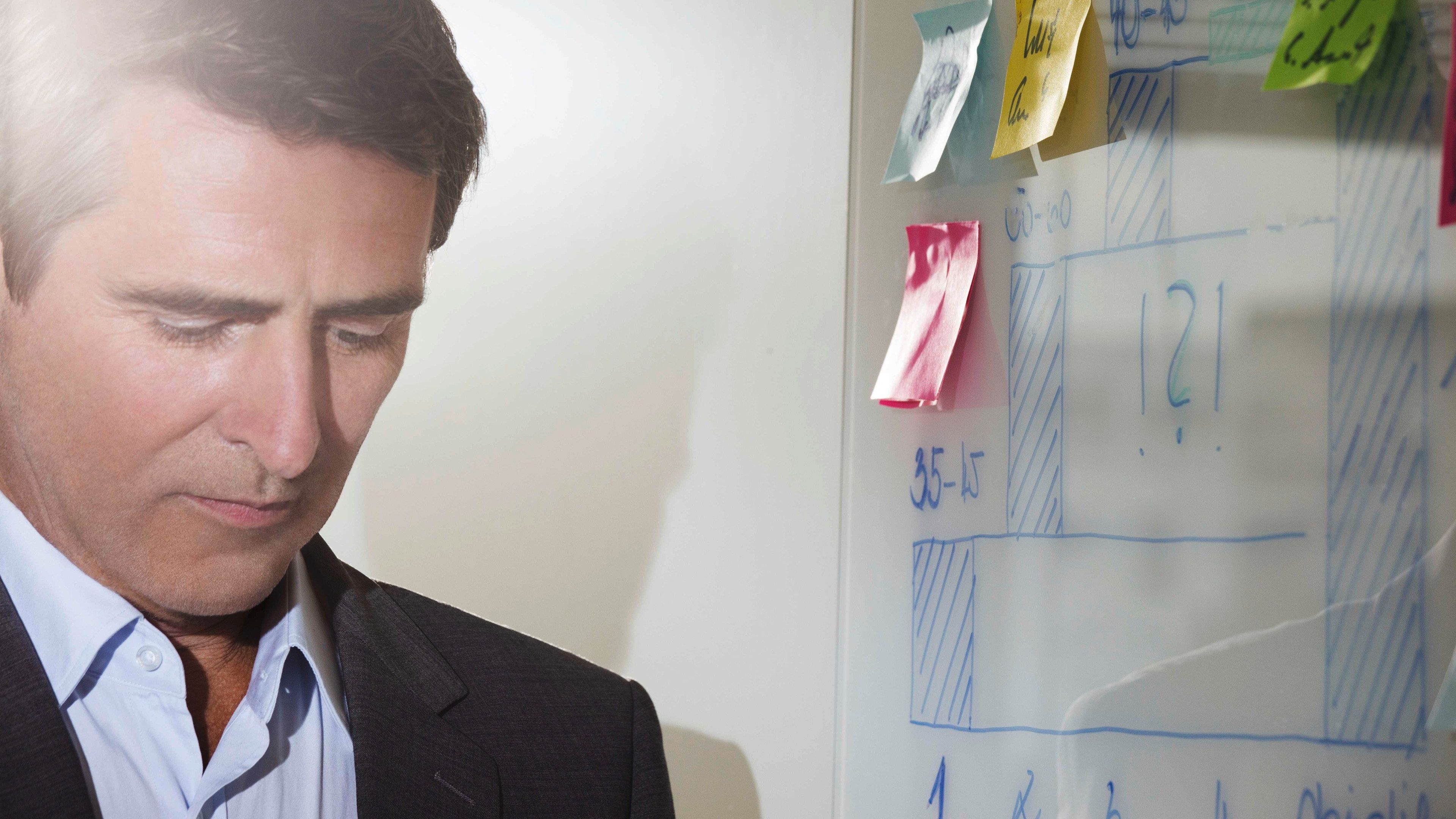 Un homme en costard est debout et à sa droite un tableau avec des post-it