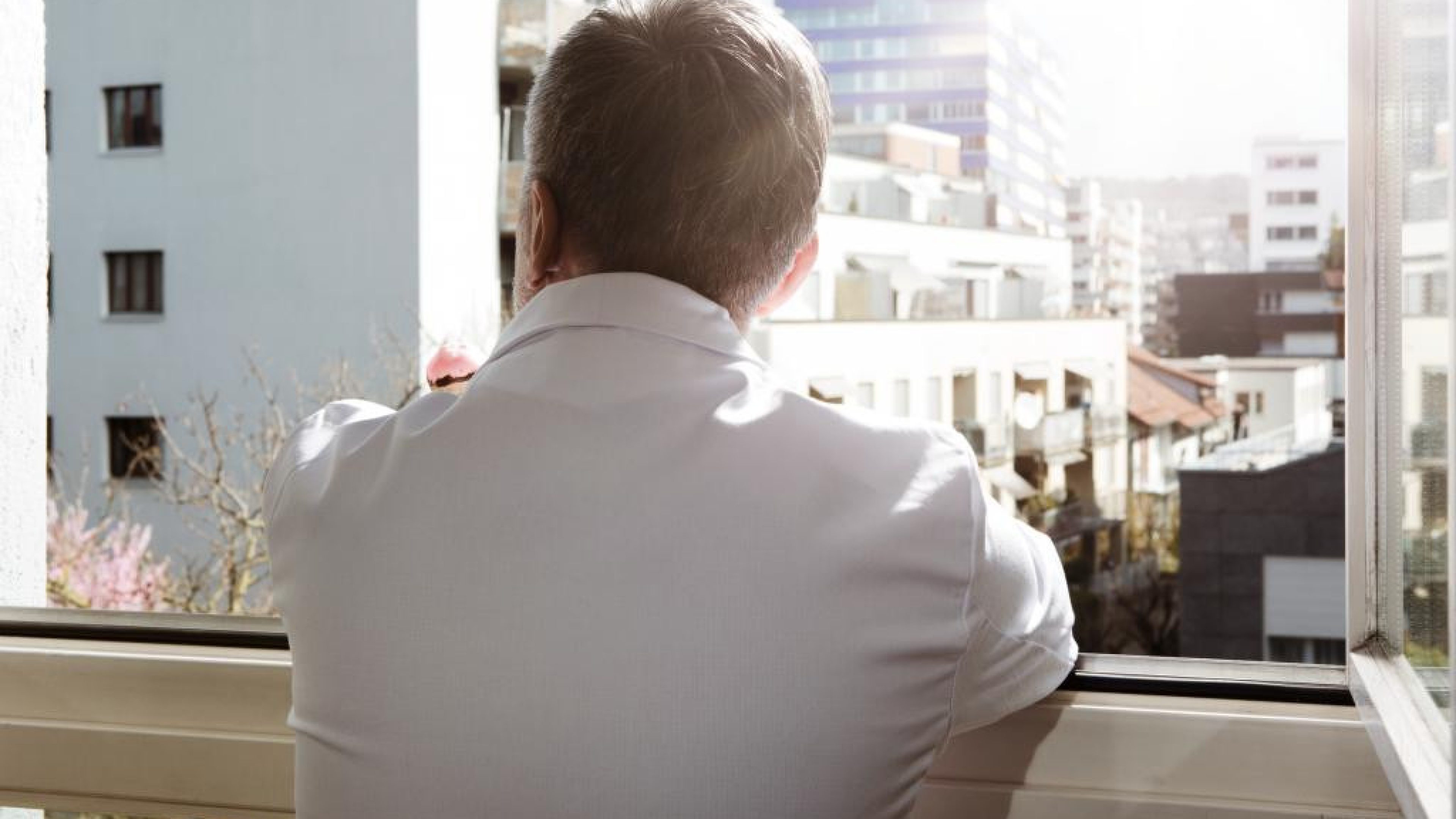 Homme en blouse blanche de dos qui regarde l'horizon par la fenêtre