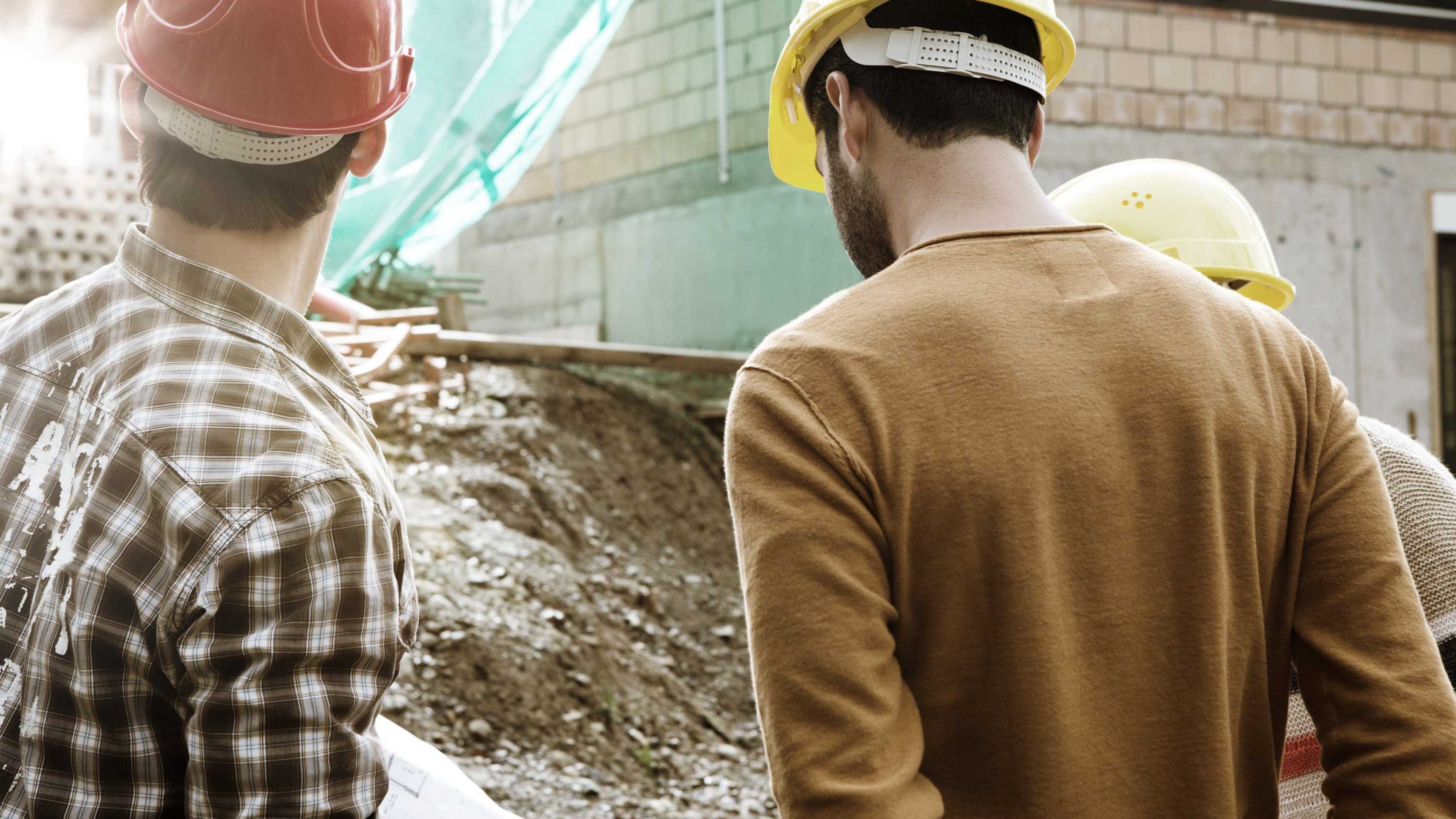 Deux personnes portant des casques de chantier observent une zone extérieure endommagée, avec un bâtiment et un tas de terre en arrière‑plan.