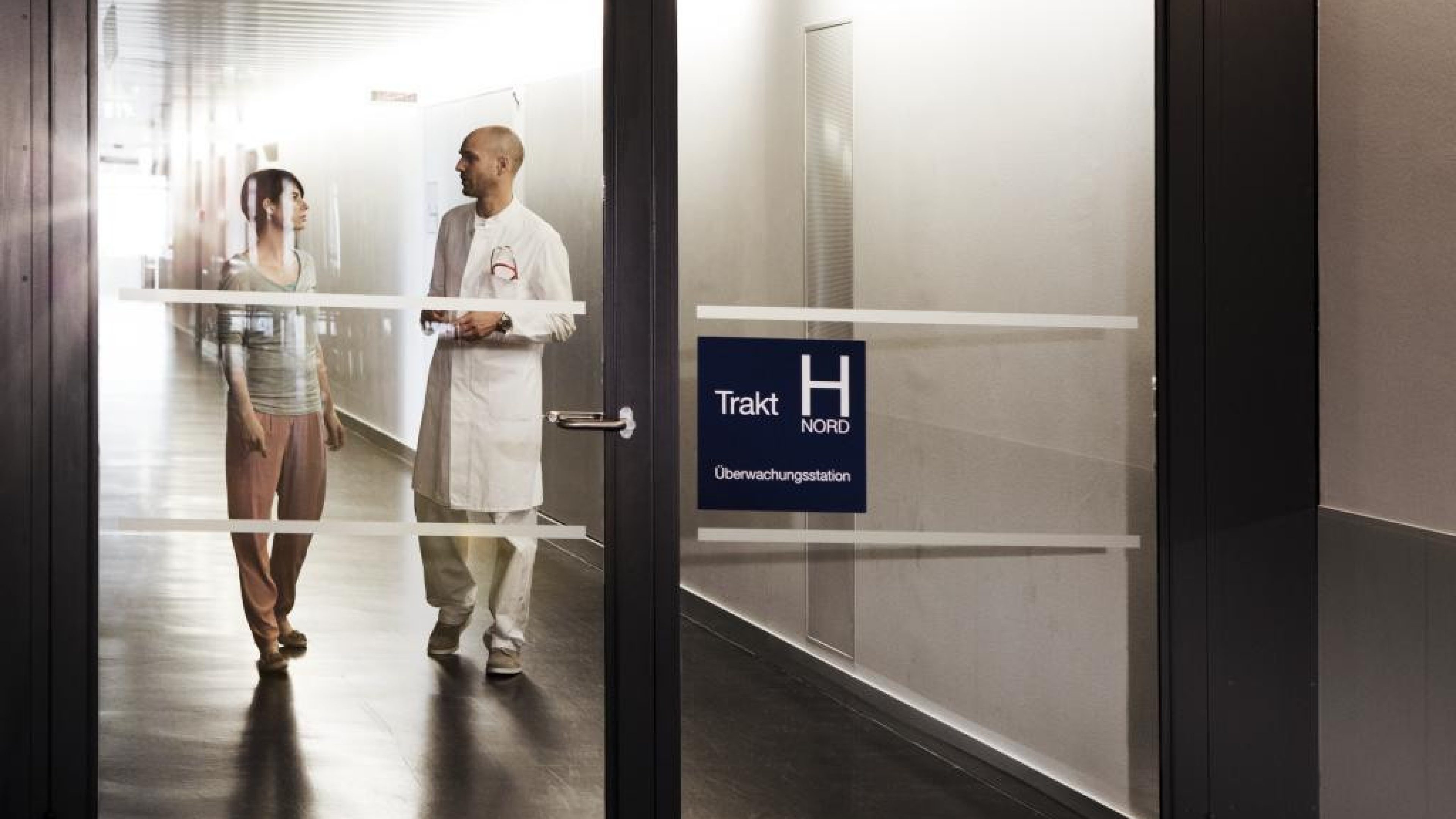 Une personne en blouse médicale échange avec une autre dans un couloir d’établissement de santé, vus à travers une porte vitrée.