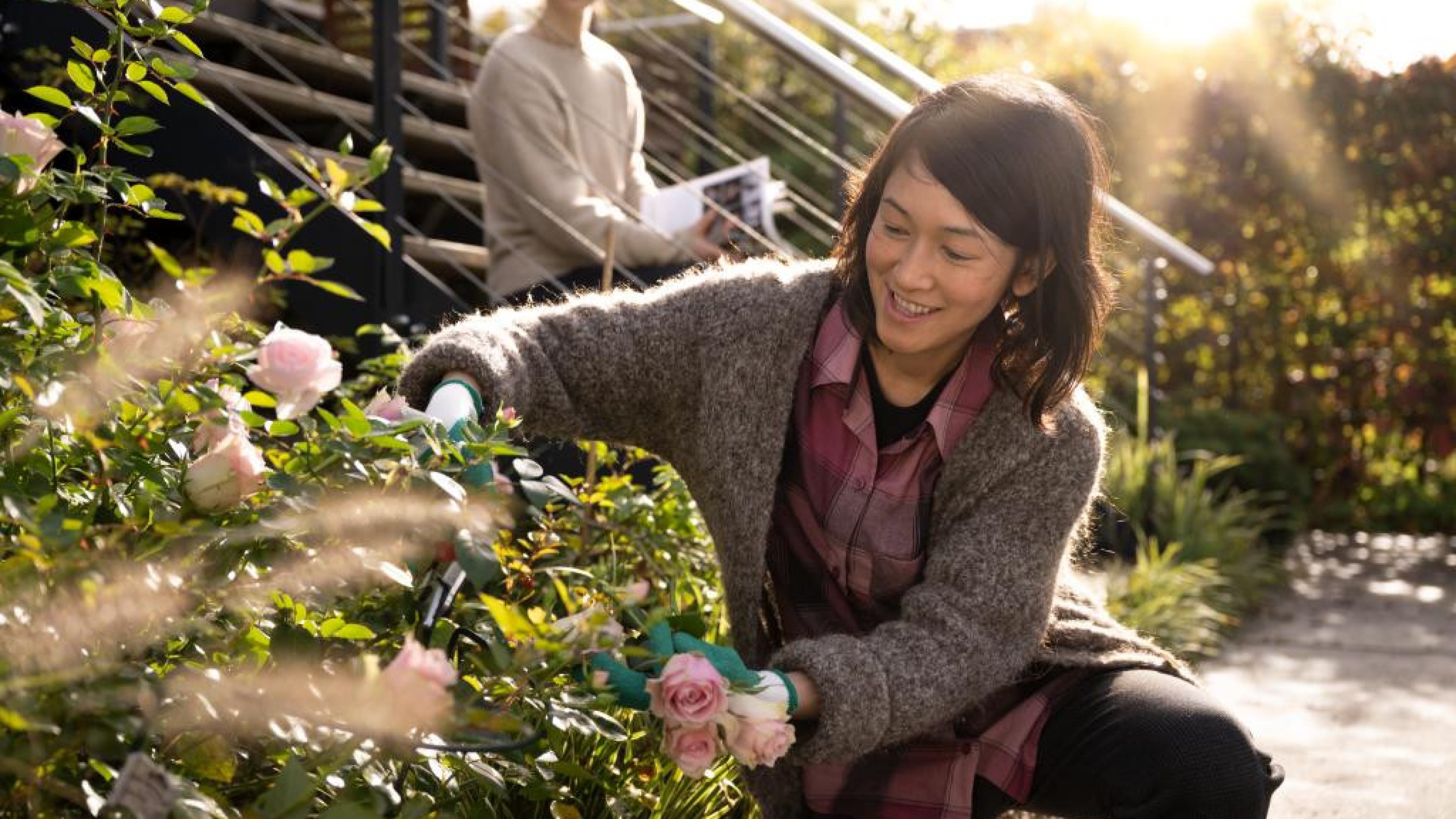 Une personne coupe des fleurs dans un jardin tandis qu’une autre se tient sur des marches à l’arrière‑plan.