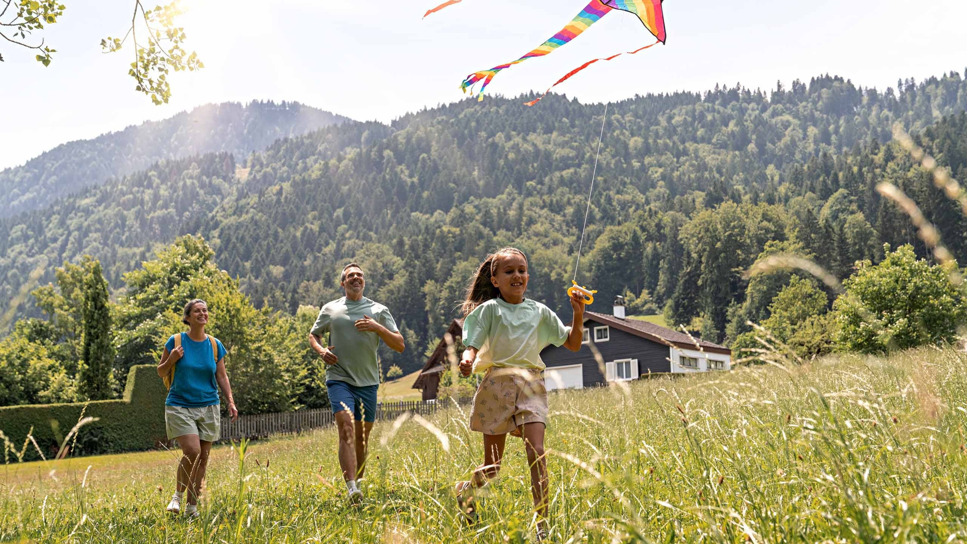 Une petite fille court dans un pré en faisant voler un cerf‑volant multicolore, accompagnée de deux adultes derrière elle, avec une maison et une forêt de montagne en arrière‑plan.