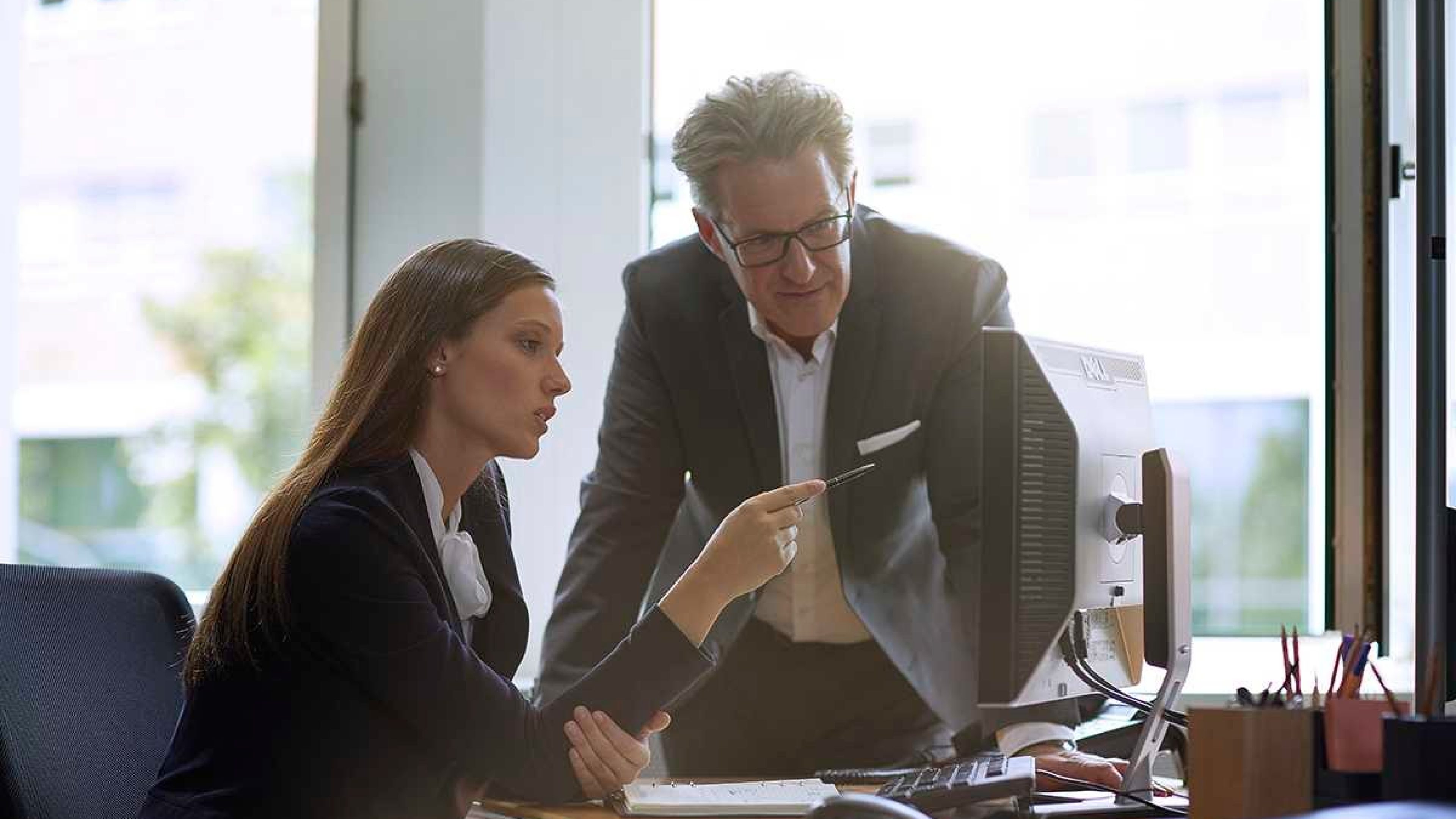 Une femme assise devant un ordinateur de bureau montre l’écran avec un stylo, tandis qu’un homme debout à ses côtés observe et l’accompagne dans la démarche.