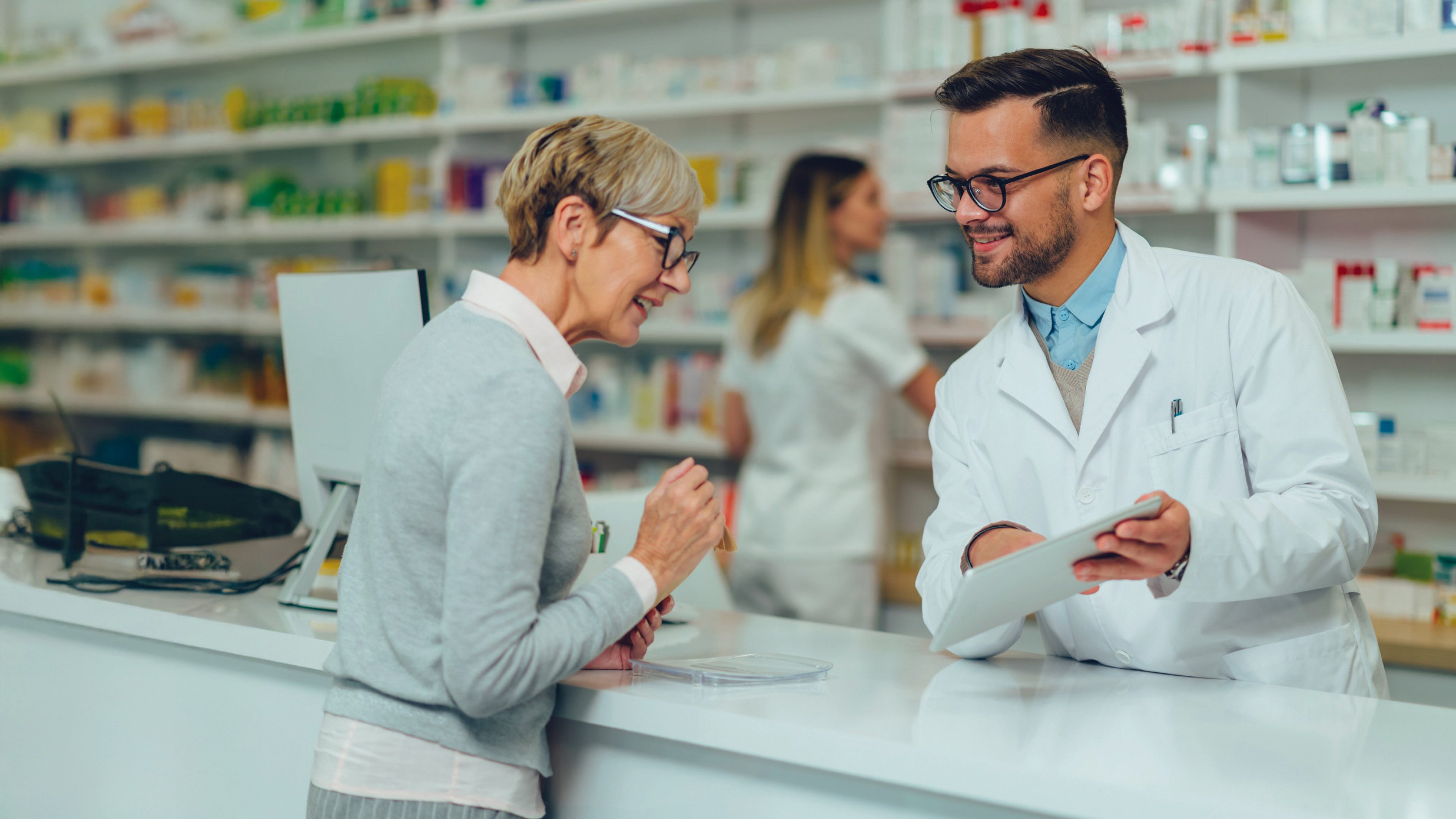 Young male pharmacist giving prescription medications to senior female customer in a pharmacy with female pharmacist in the background