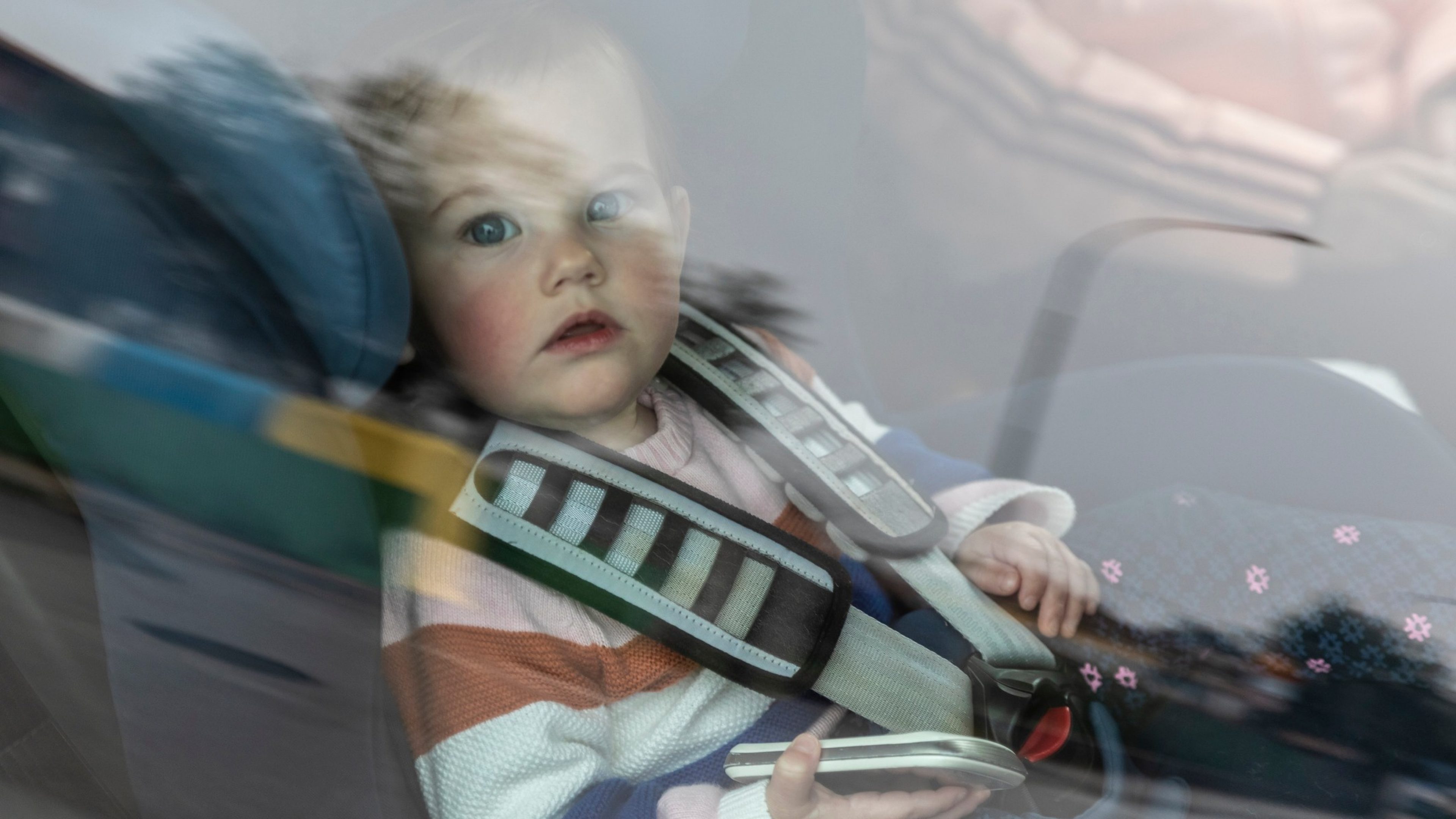 Baby girl sitting in baby car seat wearing shirt with mobile phone in hand looking outside the window worried at camera from outside.